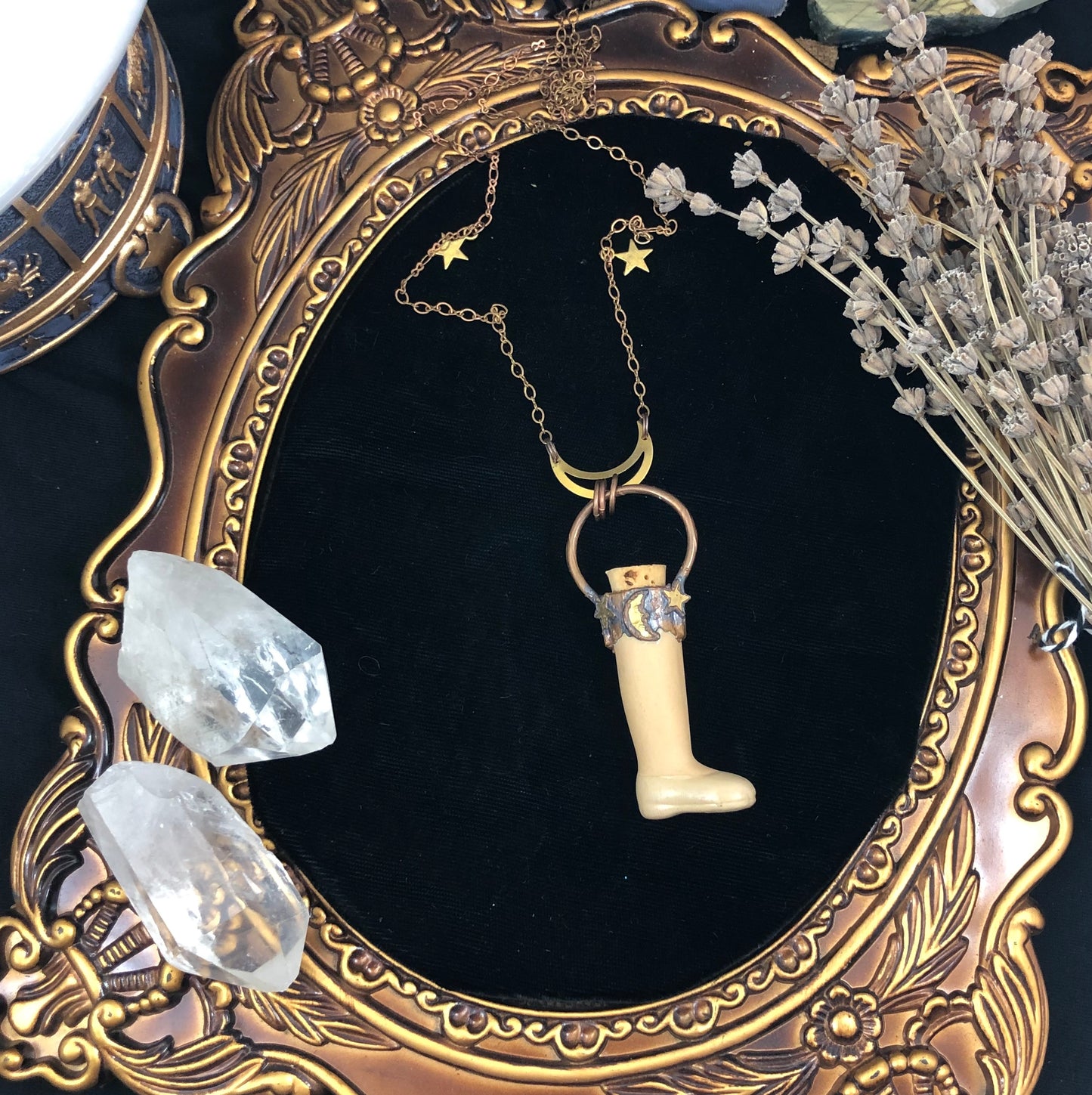A unique pendant featuring a recycled old ceramic doll leg with a copper electroformed halo ring and collar, adorned with a tiny crescent moon and two stars, displayed on a black velvet background with crystal quartz and dried flowers.