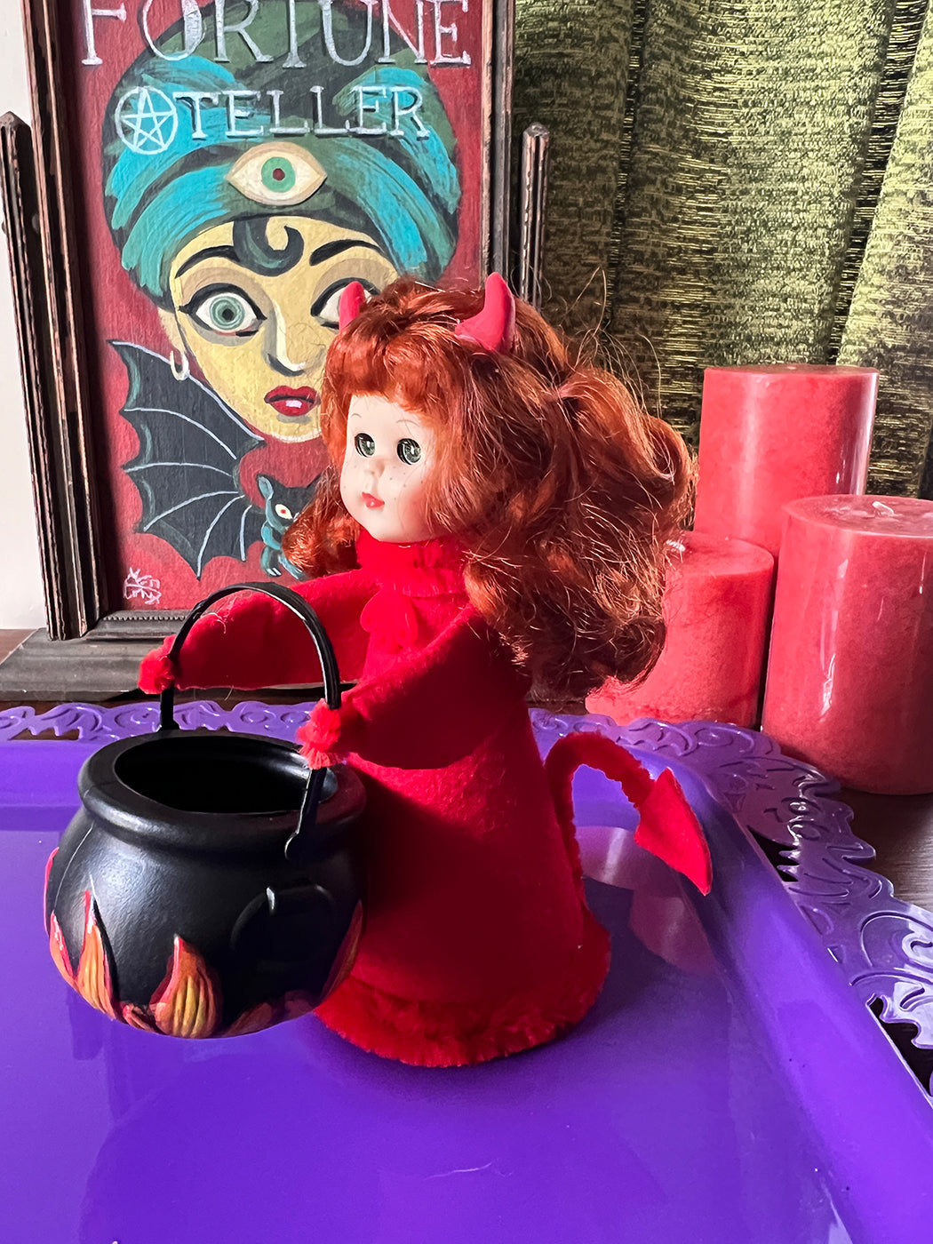 Vintage style devil doll with red outfit and black cauldron in front of a fortune teller sign.