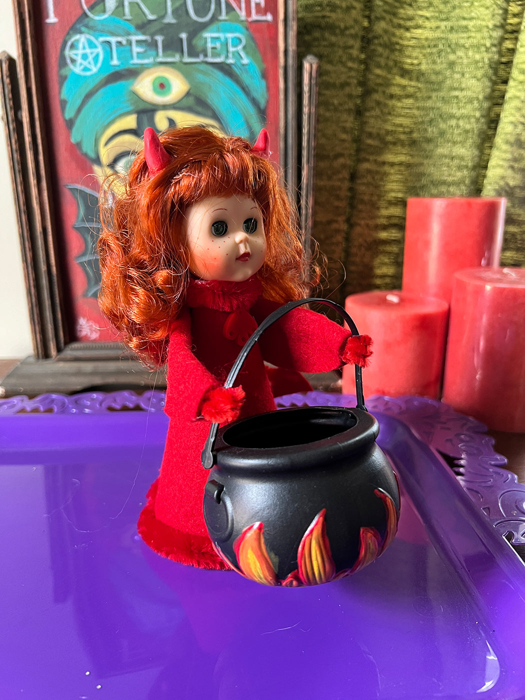 Vintage devil doll with red hair and outfit holding a black cauldron on a purple surface.