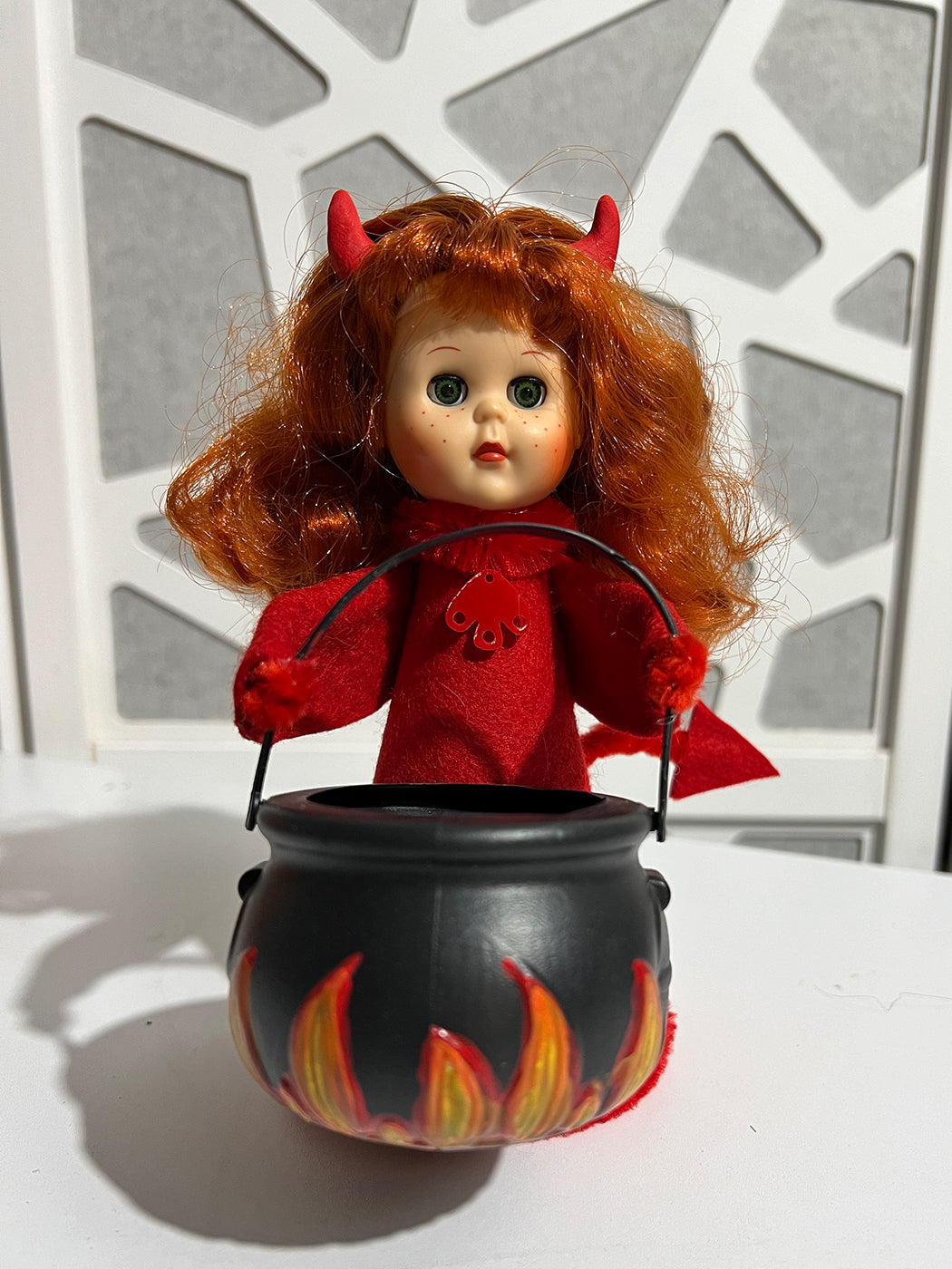 Devil-themed doll with red hair and horns holding a black cauldron with flames against a white lattice background.