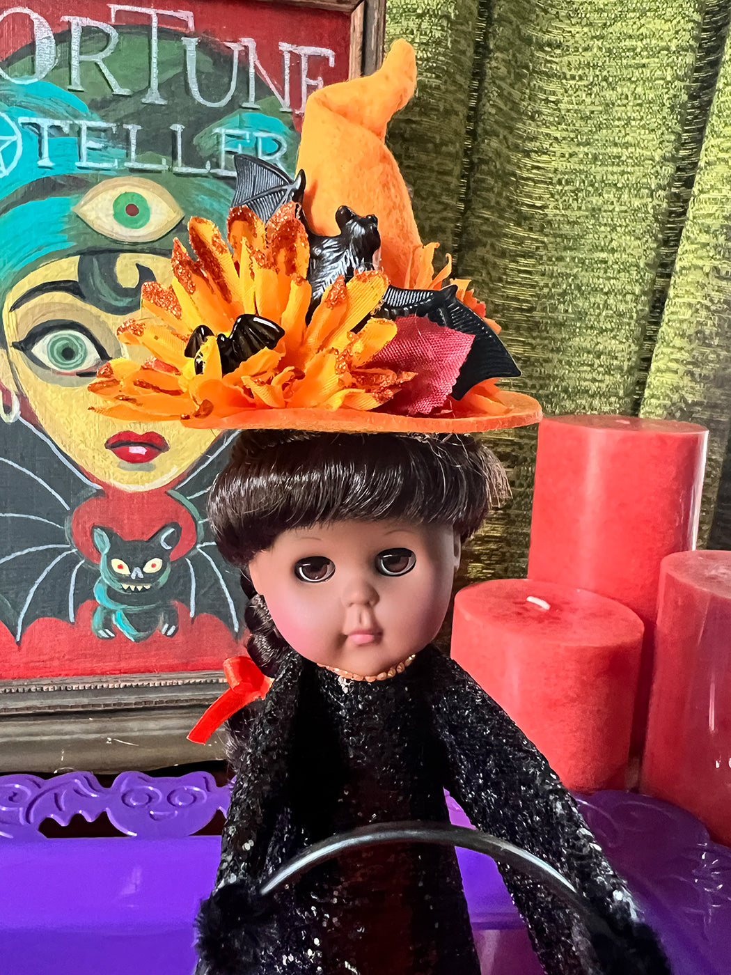 Doll with a witch hat and black outfit in front of a colorful painting and red candles.