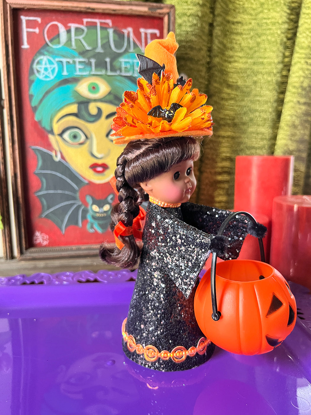 Glamorous witch doll with black glitter dress, orange hat, and pumpkin bucket in front of a colorful poster.
