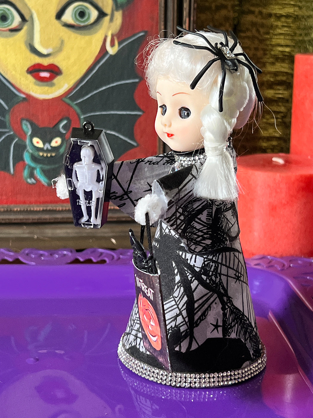Vintage-style doll with black and white outfit and spider decoration, with a painting and red candle in the background.