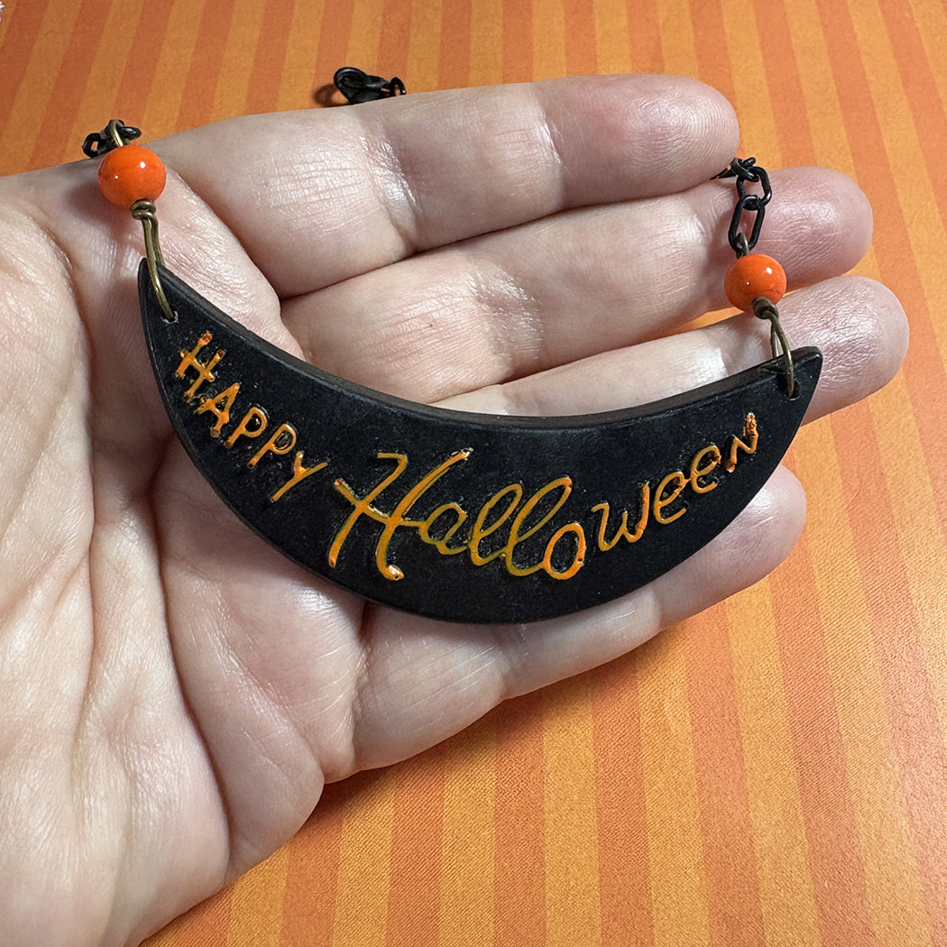 Black 'Happy Halloween' crescent moon ornament held in a hand against an orange background
