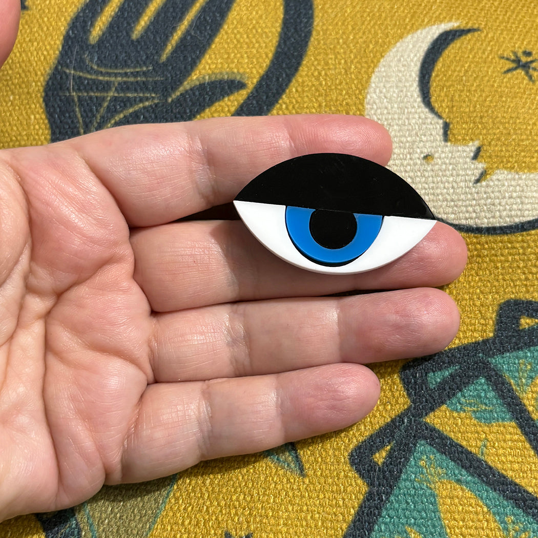 Hand holding a black and white eye brooch with a blue center against a patterned background