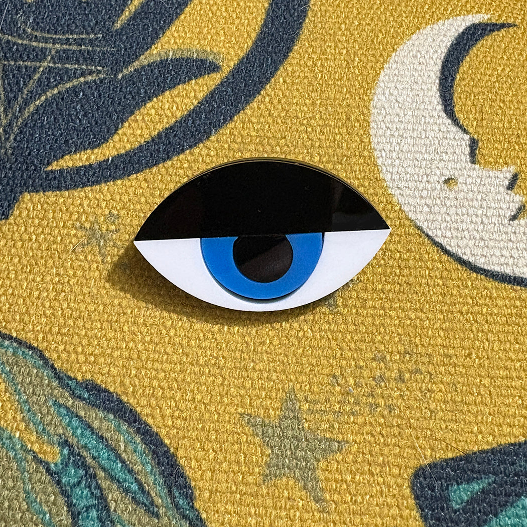 Modernist eye pin with blue center on a textured yellow background with moon and star designs.