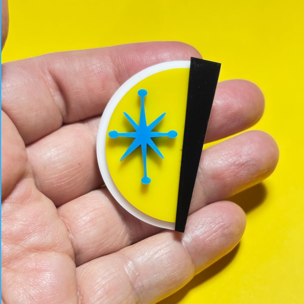 A hand holding a small oval pin or badge with a bright yellow center, a blue starburst-like symbol, a white border, and a black vertical strip on a bright yellow background.