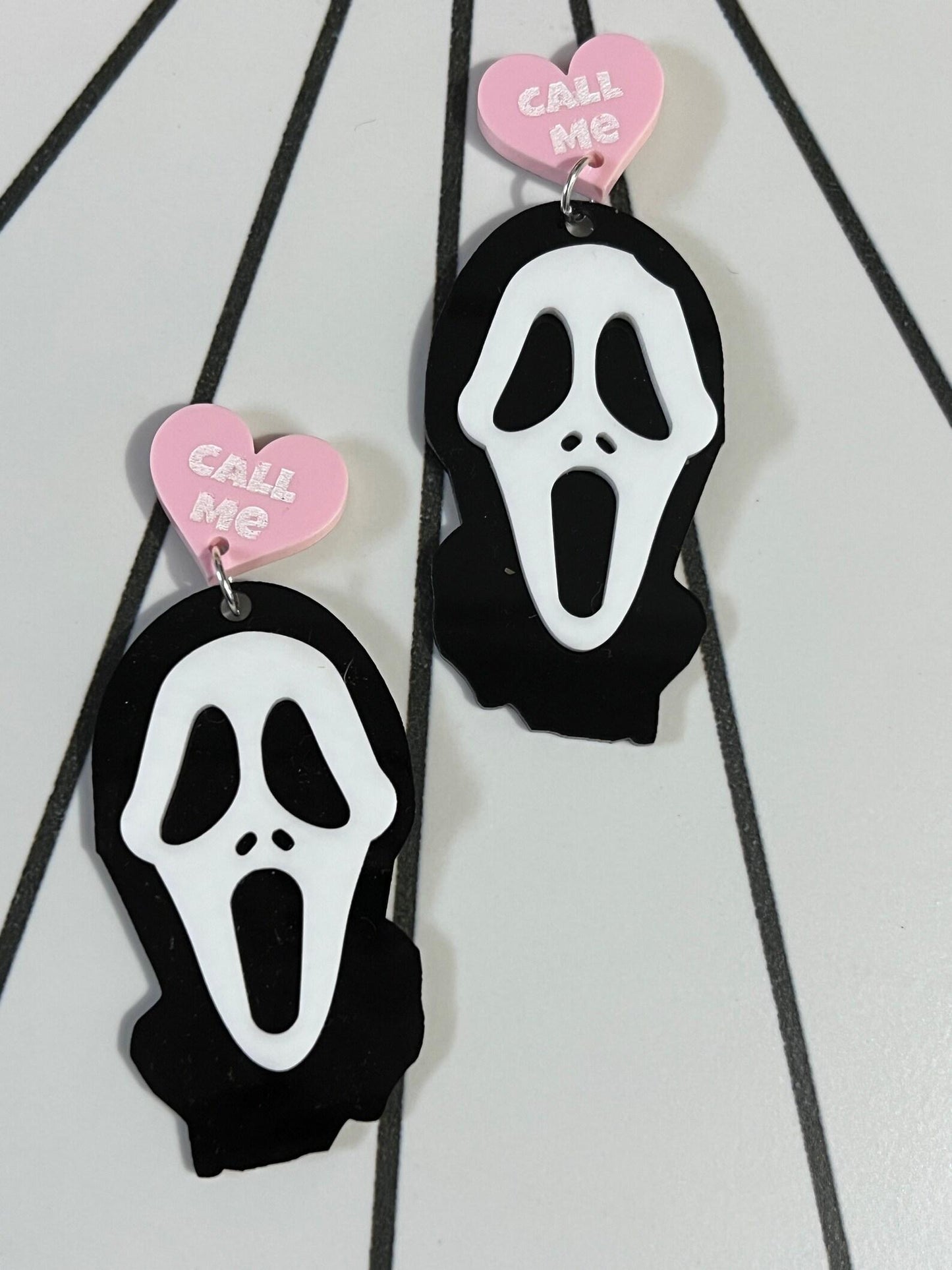 Earrings with ghost face design and 'Call Me' heart tag on a striped background