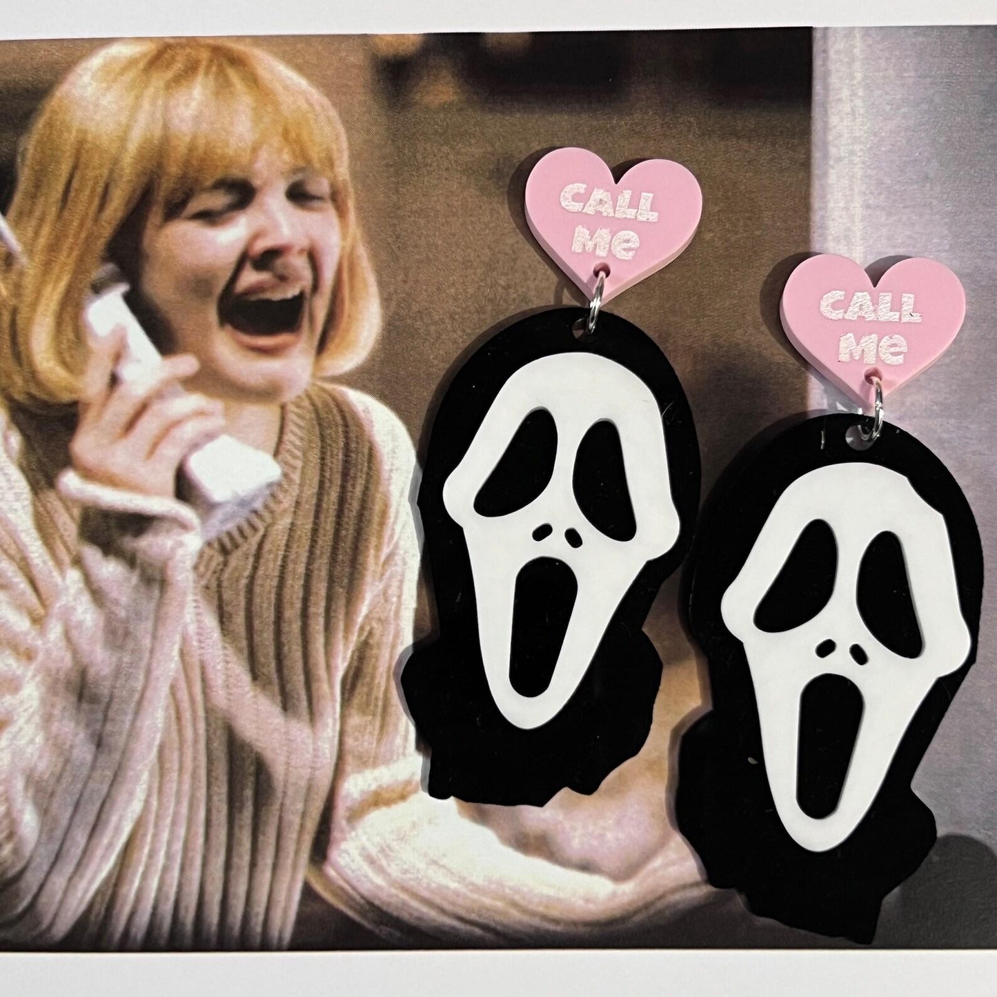Scream ghostface earrings with 'Call Me' heart tags on a background of a woman on the phone.