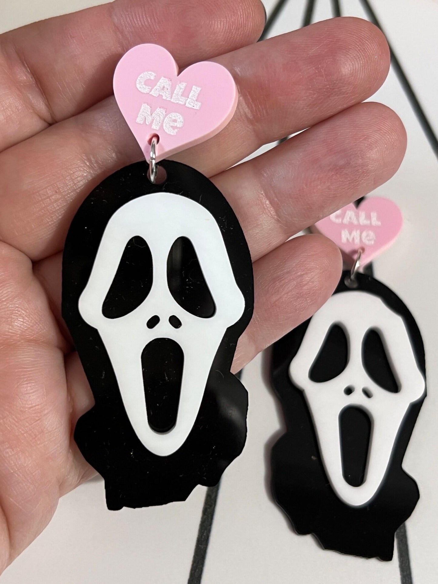 Earrings with ghost face design and pink heart-shaped tag on a hand.
