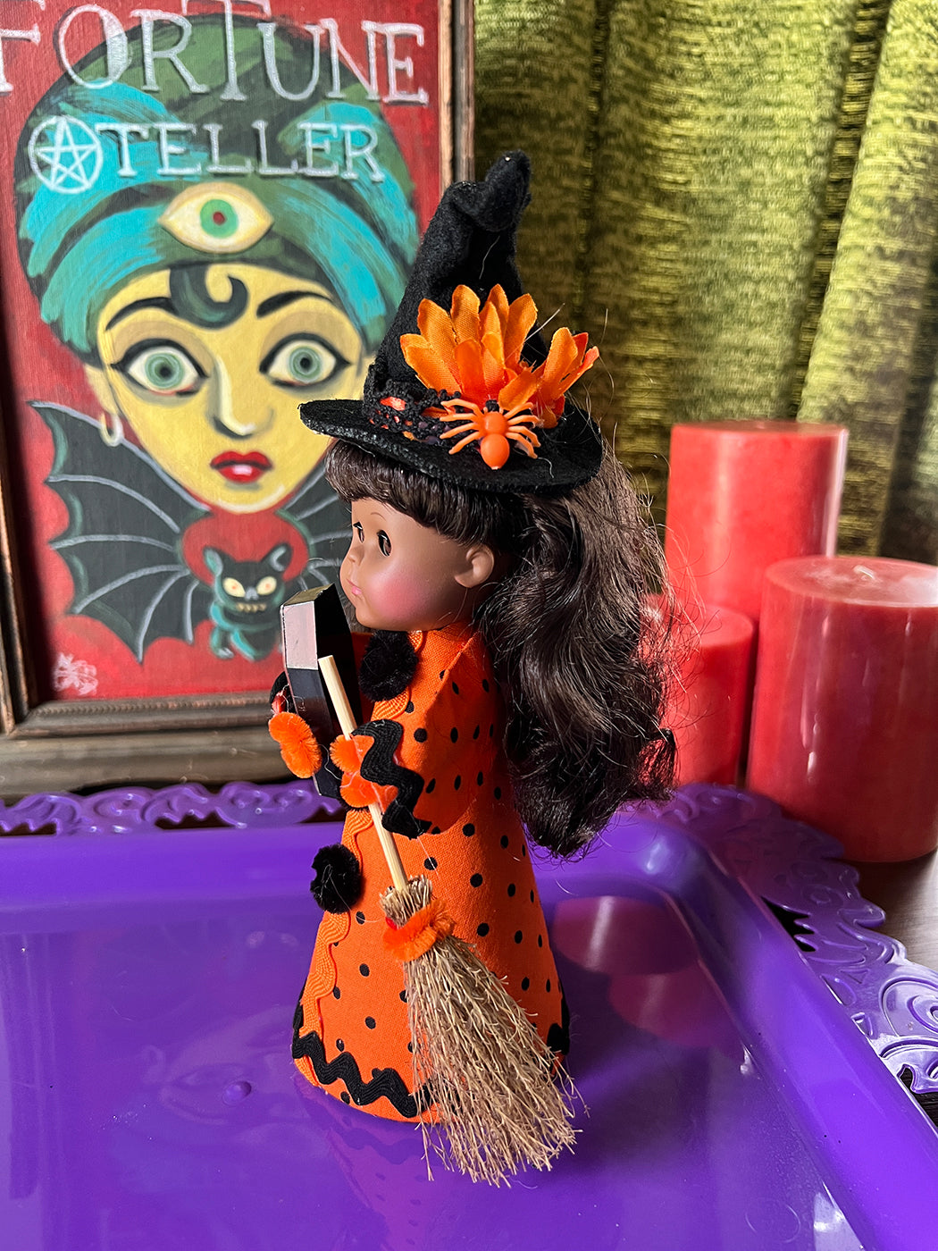 Halloween-themed doll in an orange outfit with a broom, surrounded by candles and a fortune teller painting.