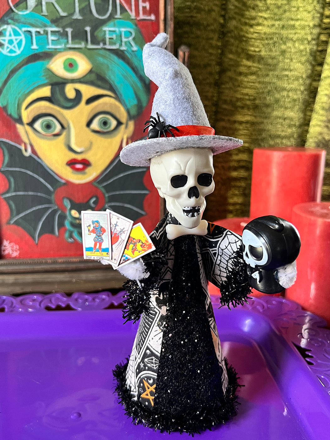 Skull head figure in a witch costume with tarot cards, surrounded by colorful decor