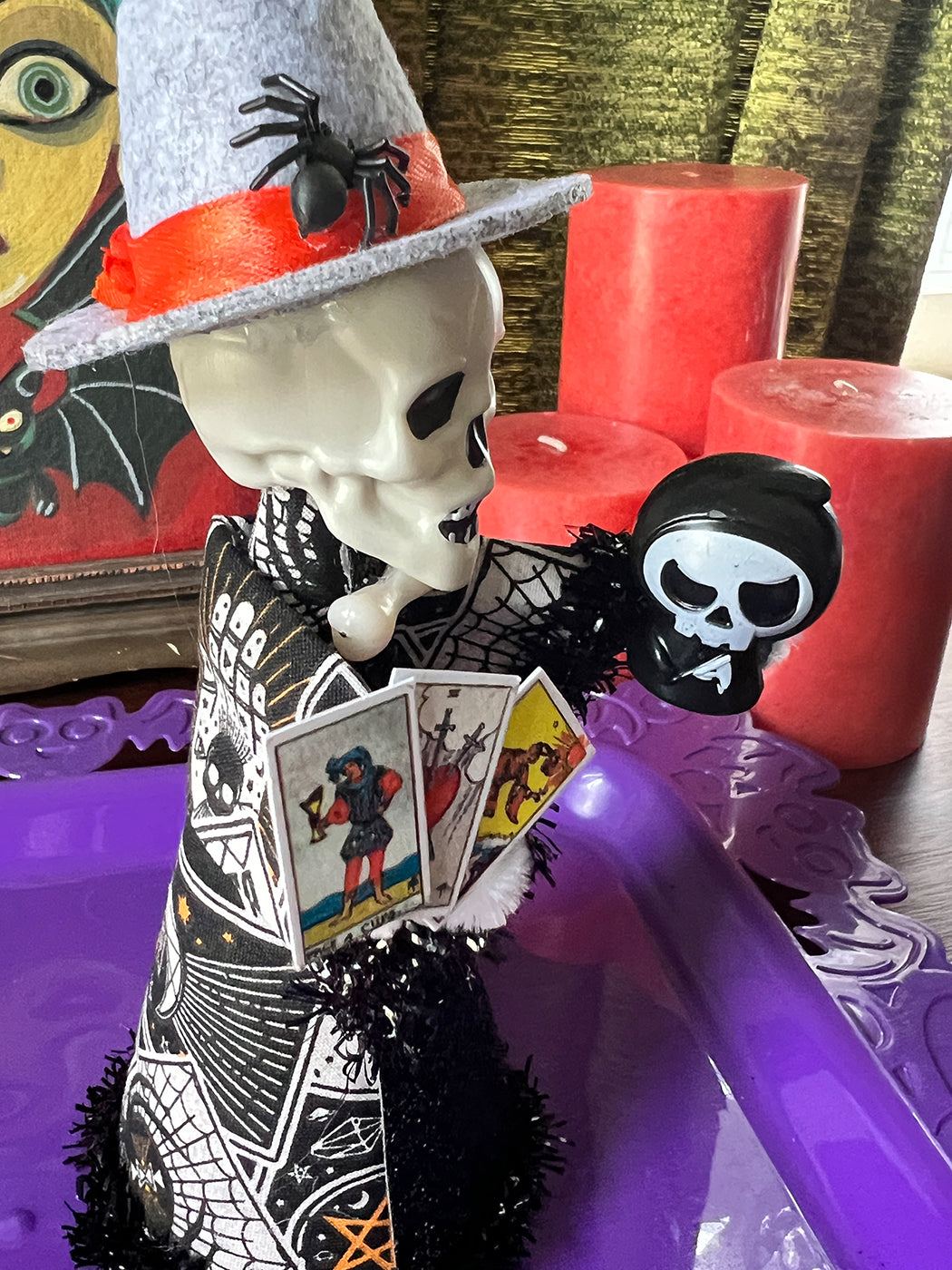 Decorative skeleton figure with a hat, tarot cards, and candles on a purple surface.