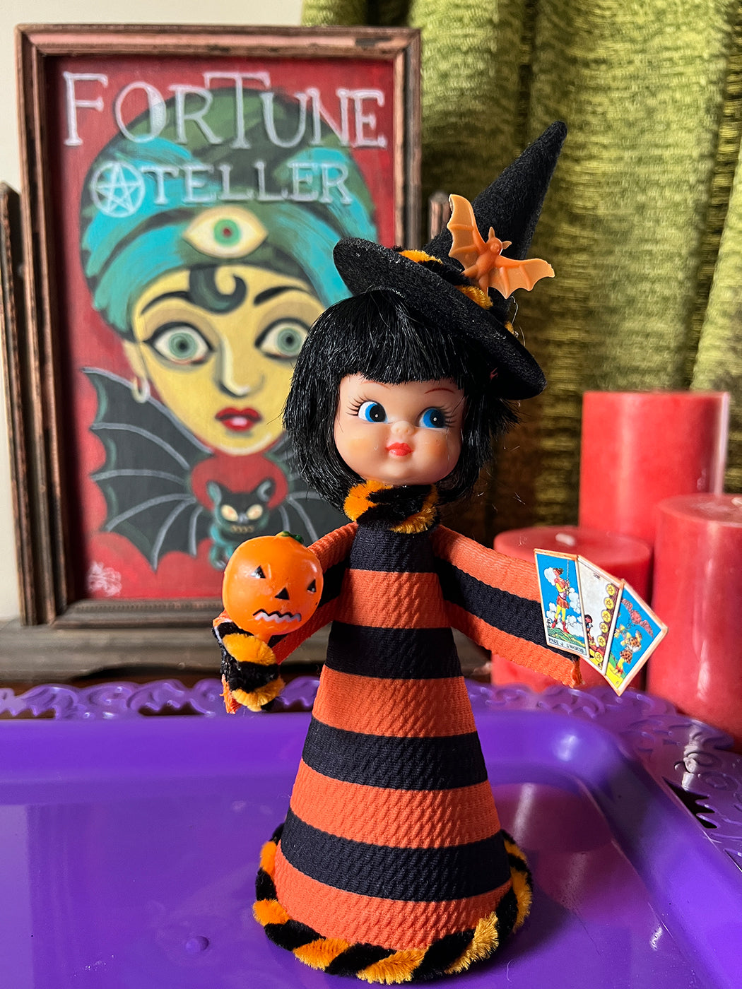 Halloween-themed doll in a black and orange outfit with a pumpkin, surrounded by candles and a framed picture of a fortune teller.