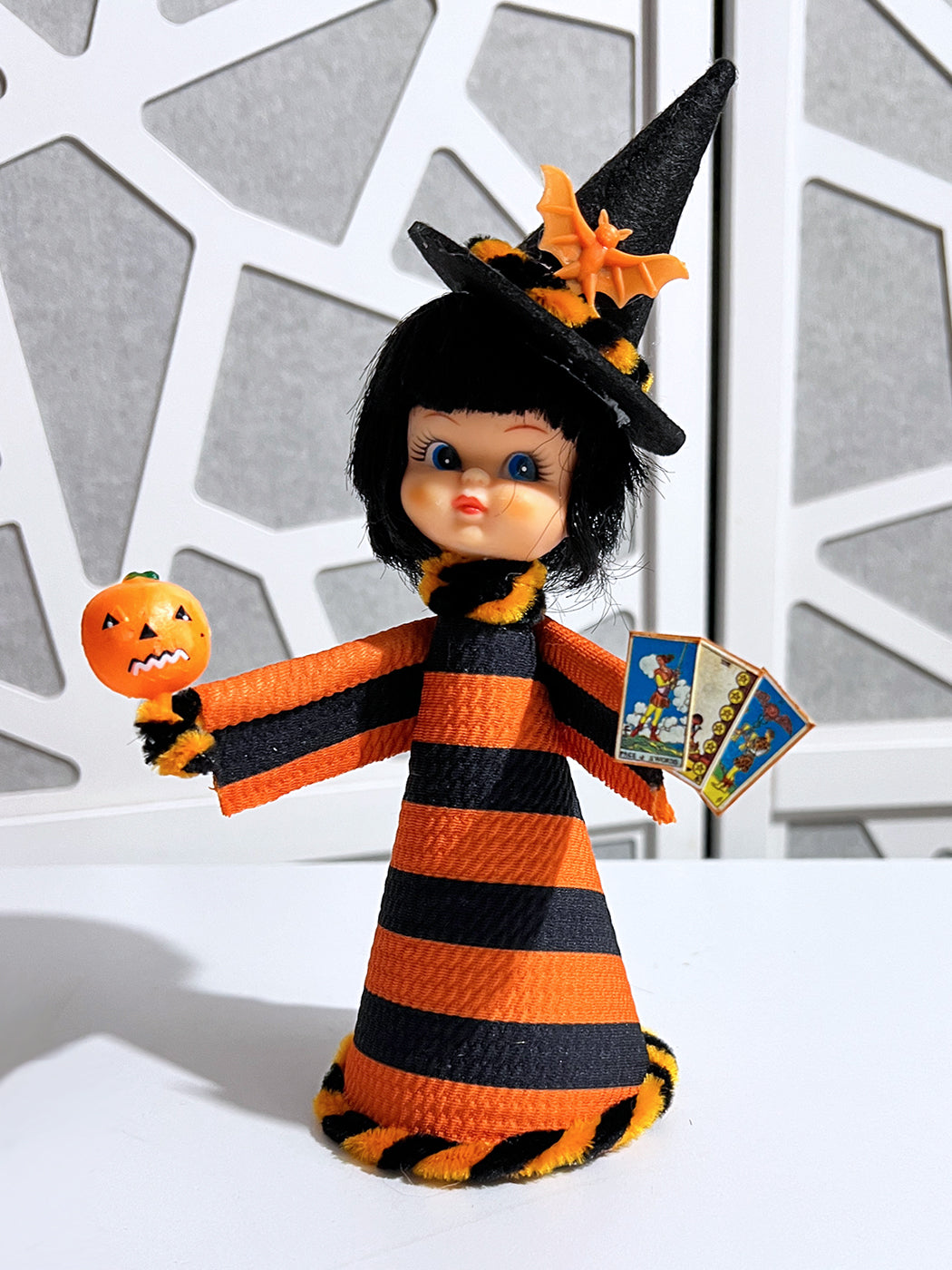 Halloween-themed doll with black and orange outfit, pumpkin, and cards against a geometric-patterned wall.