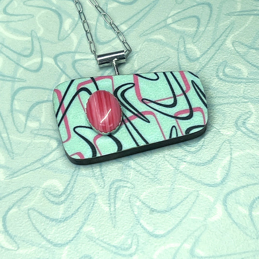 A colorful geometric atomic boomerang patterned necklace with a vibrant pink Rhodochrosite stone set in a sterling silver pendant on an aqua-colored background.