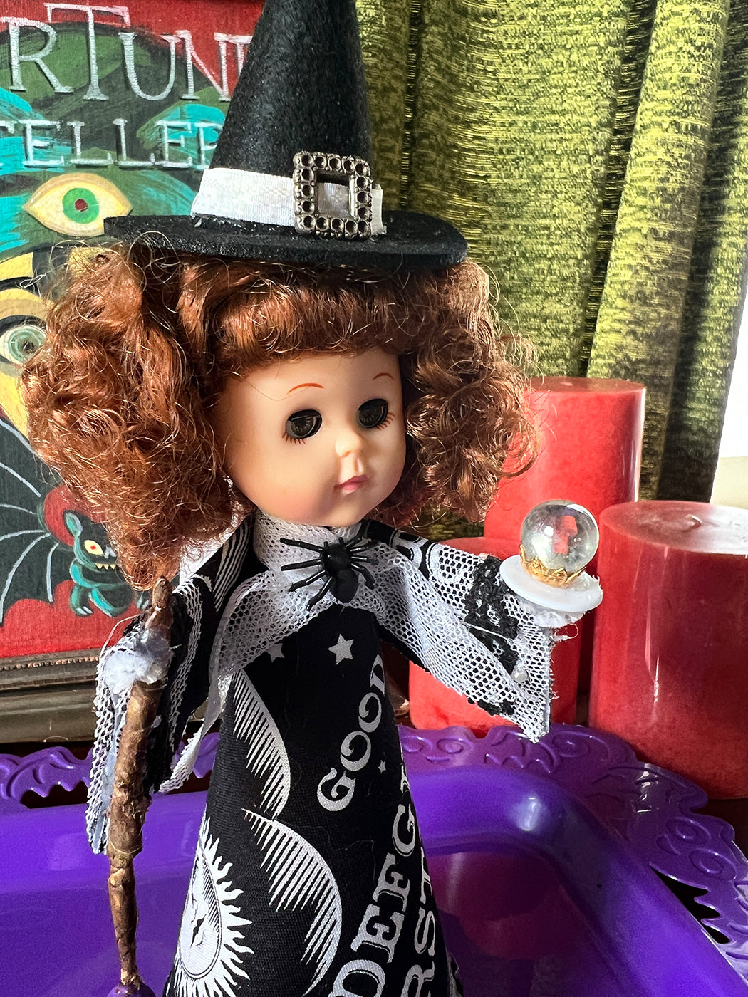 Vintage-style witch doll with a black hat and cape, holding a crystal ball, on a purple surface.