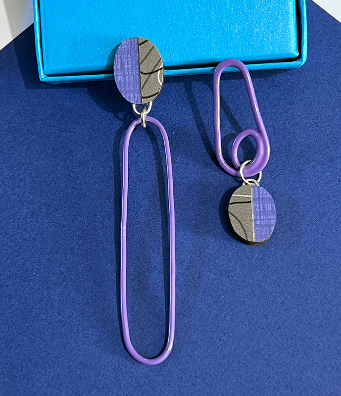 Close of of asymetrical purple and gray laminate and powdercoated earrings on blue background.