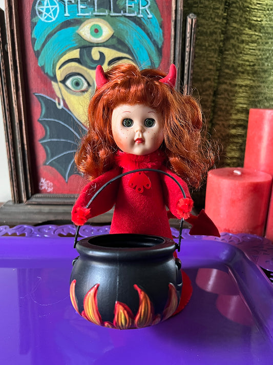 Vintage-style Devil doll with red hair and outfit, holding a black cauldron with flames on a purple surface.