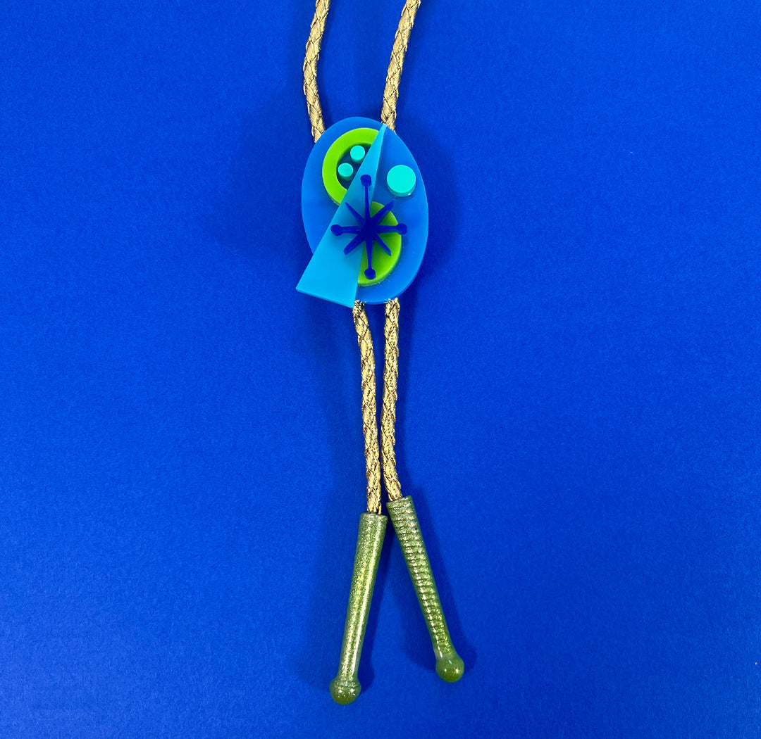 A bold 60s inspired mid century modern bolo tie in blues and green on a gold vegan leather cord with green tips. Retro bolo tie on blue background.