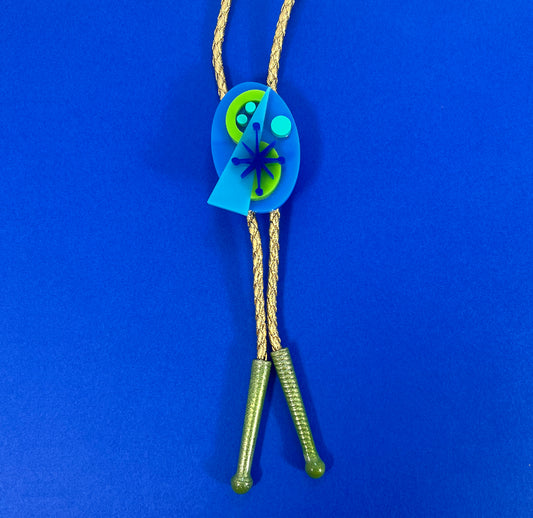 A bold 60s inspired mid century modern bolo tie in blues and green on a gold vegan leather cord with green tips. Retro bolo tie on blue background.