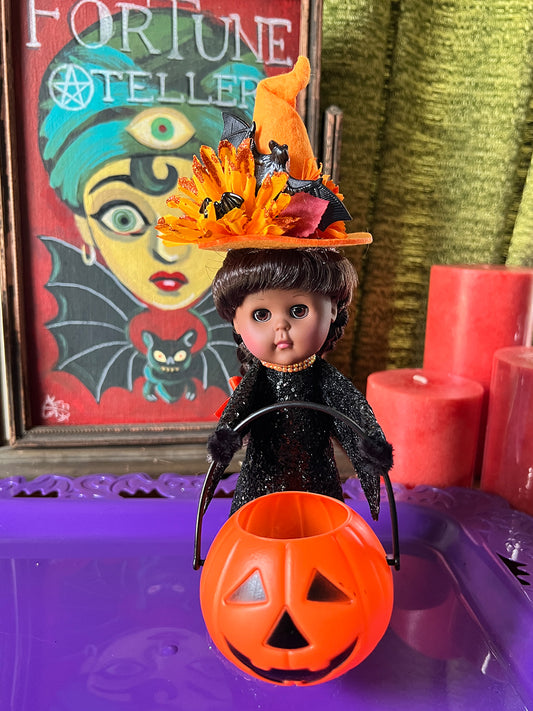 Halloween-themed doll with a jack-o'-lantern bucket and decorative hat in front of a colorful painting.