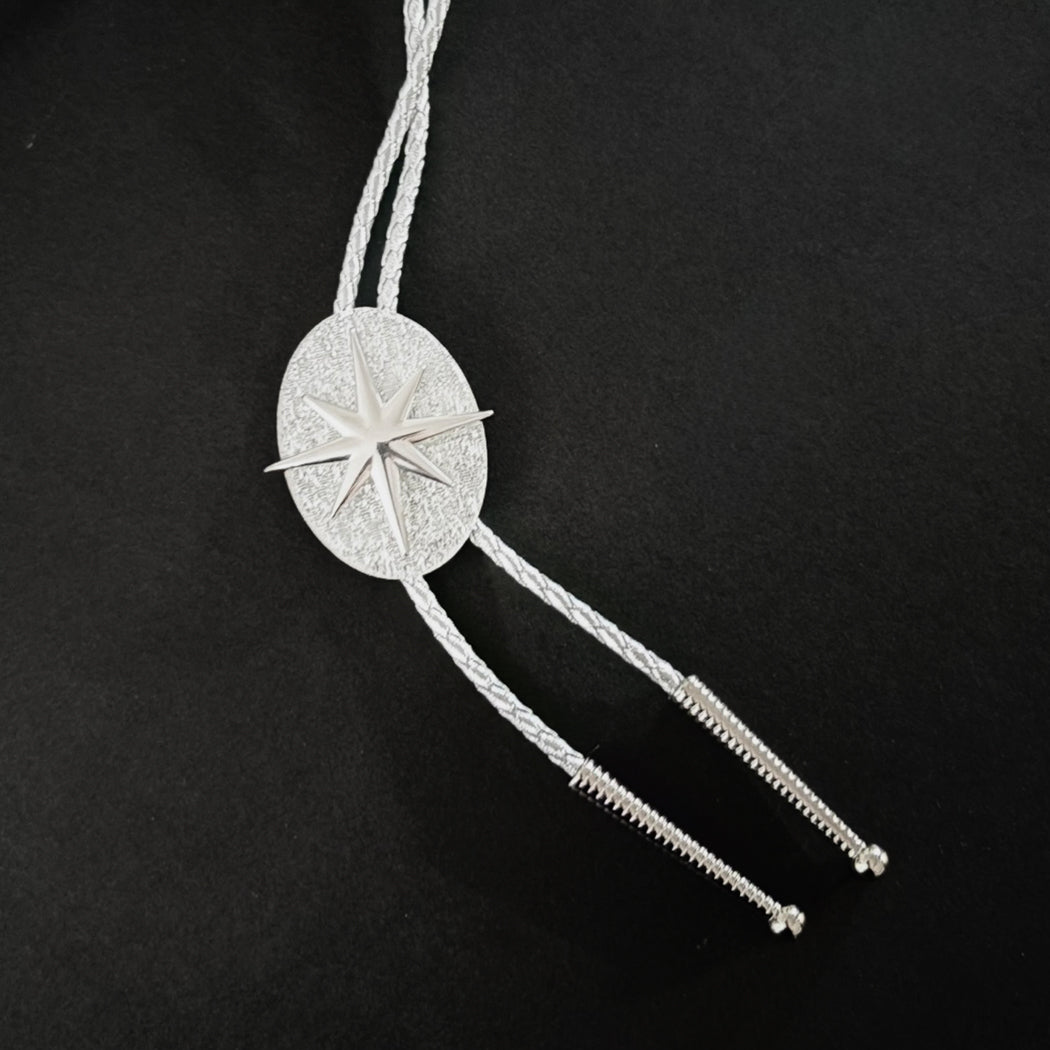 A silver-toned bolo tie featuring a textured oval center with a starburst motif and braided cord tails, displayed on a solid black background.