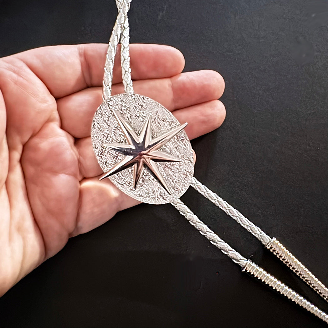 A hand holding a silver-toned western bolo tie with a textured oval medallion and starburst center, against a dark background.