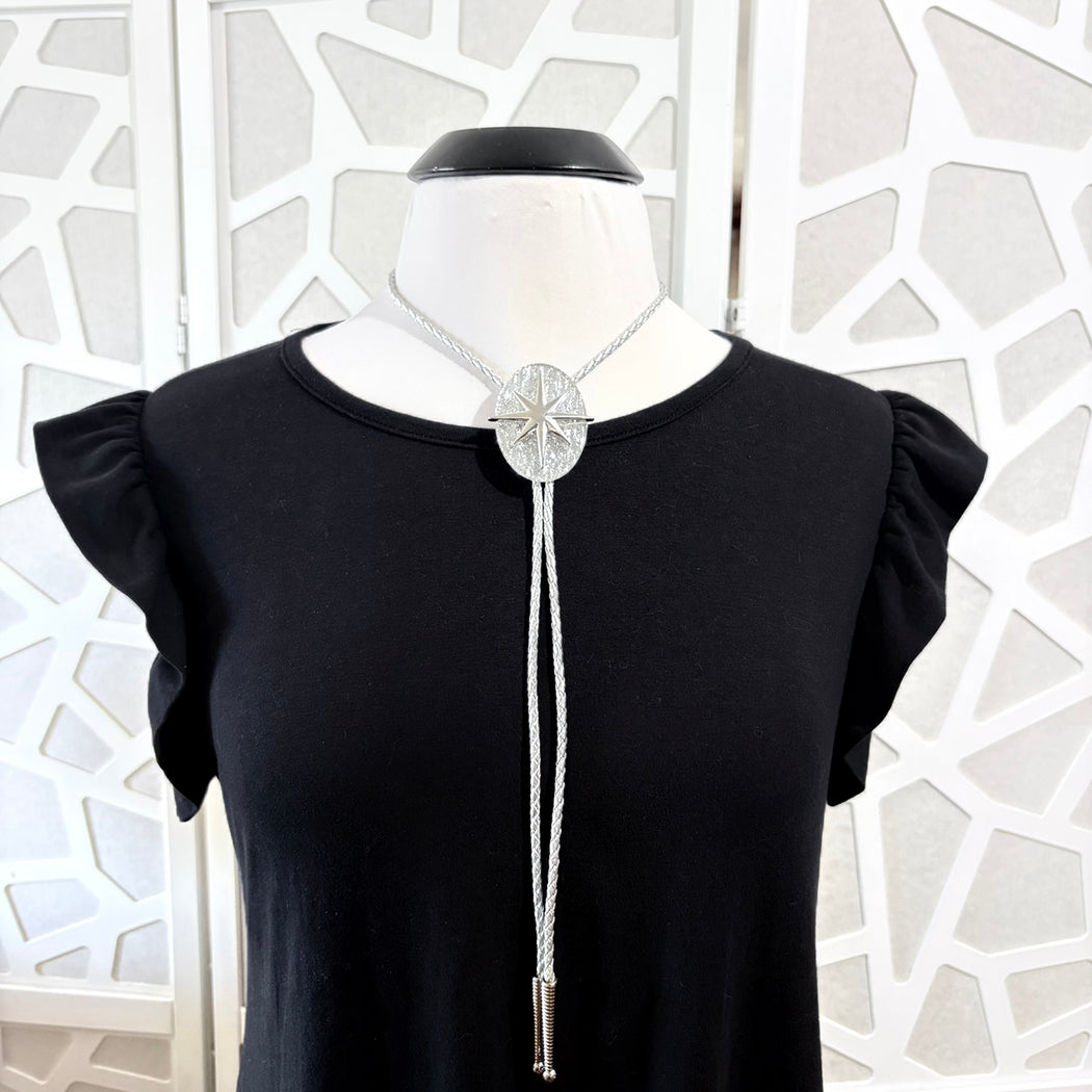 A silver-toned bolo tie necklace with a starburst center and braided cord, displayed on a mannequin wearing a black top. The mannequin is set against a white folding screen with a geometric pattern.