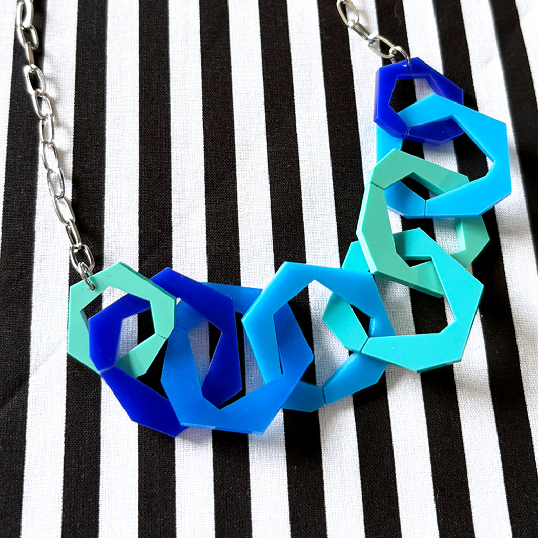 Geometric necklace with blues and aqua links on a black and white striped background