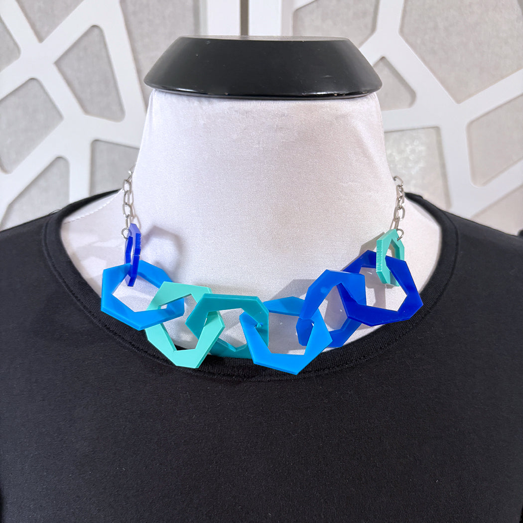 Necklace with blue and aqua geometric links on a mannequin against a patterned wall.