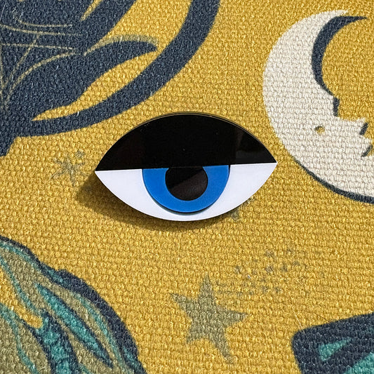 Modernist eye pin with blue center on a textured yellow background with moon and star designs. 