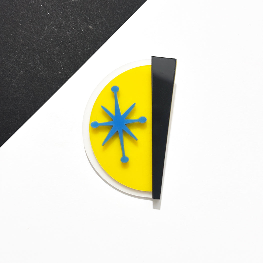 A modern semi-circular pin or badge with a bright yellow left half, a black right half, and a blue six-pointed starburst in the center, photographed on a white surface with a black diagonal background section.