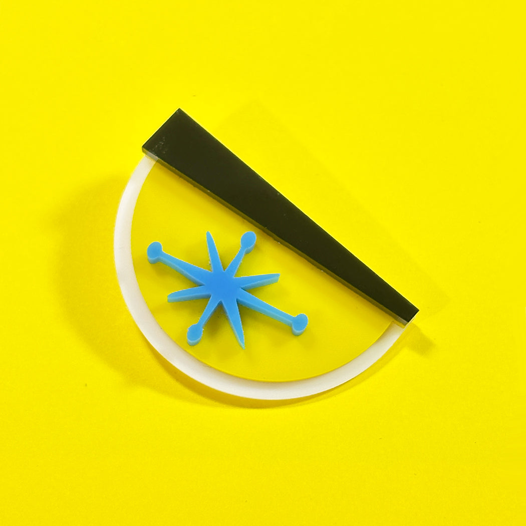 A retro-style semi-circular acrylic pin with a blue starburst center, a white curved border, and a black diagonal bar, set against a solid bright yellow background.