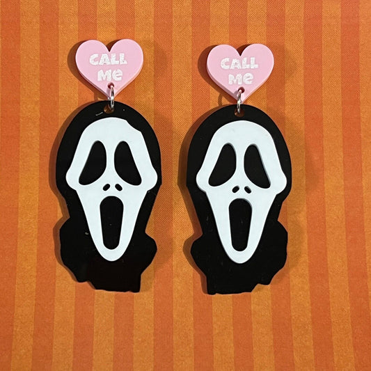 Pair of ghost face earrings with pink hearts and 'Call Me' text on an orange striped background.