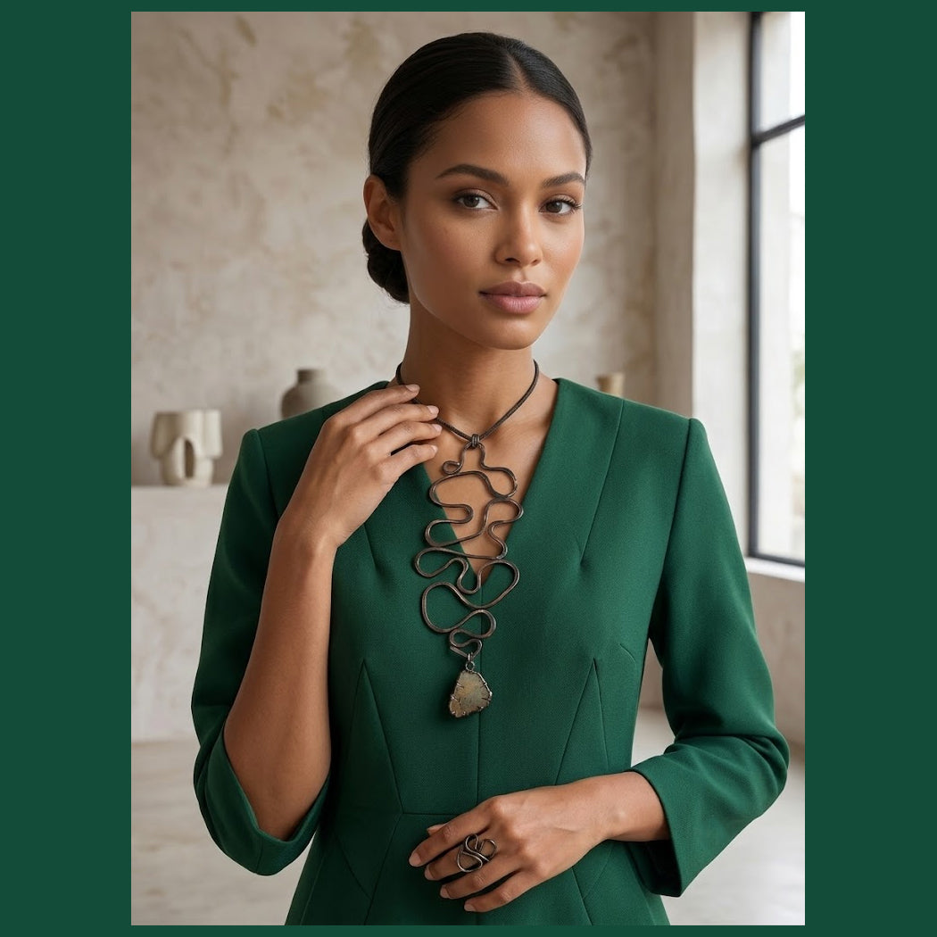 Dark haired woman in a green dress wearing a large copper glyph necklace in front of pottery on a shelf.

