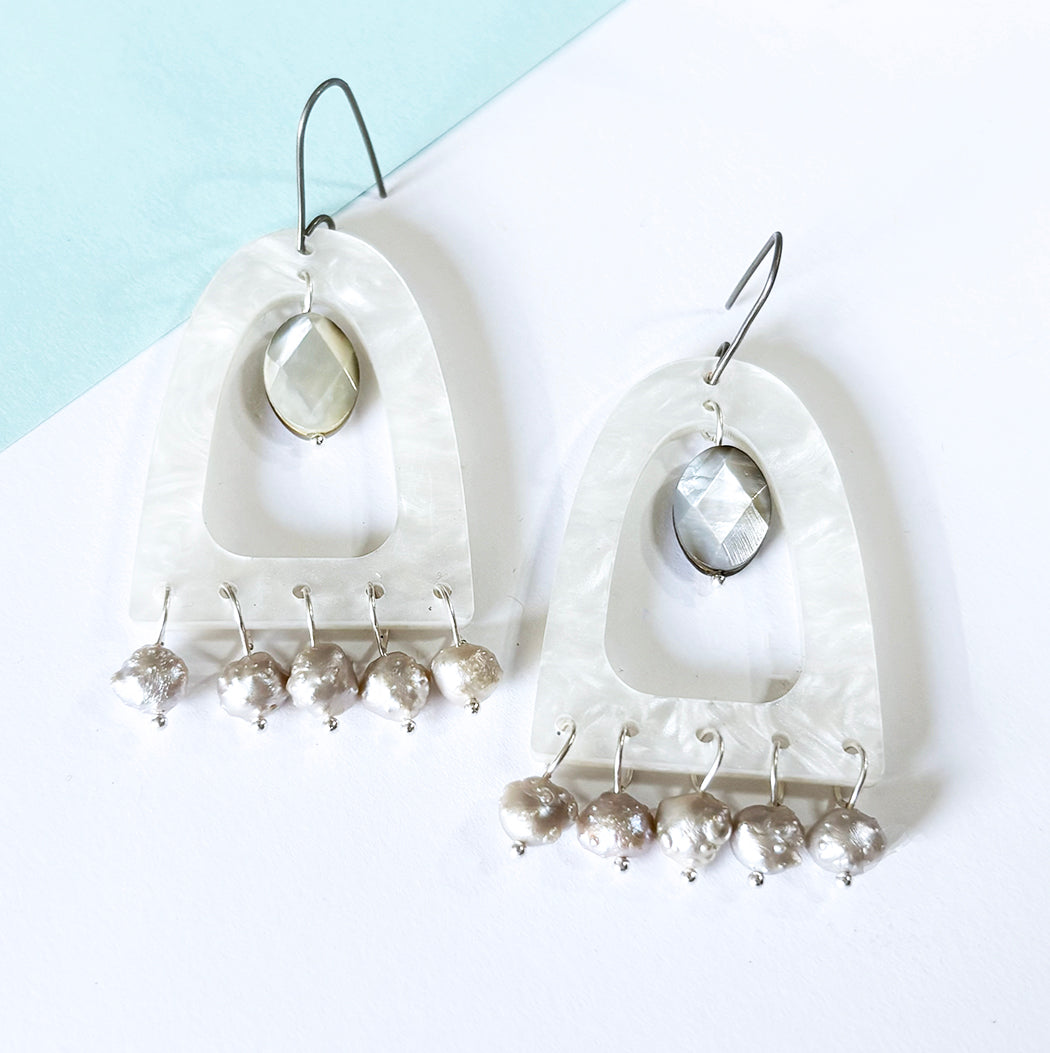 A pair of bohemian dangle earrings featuring milky white mother-of-pearl acrylic arches, mother of pearl facetted beads and dangling silver baroque druzy pearl beads, displayed on a white and light blue background.