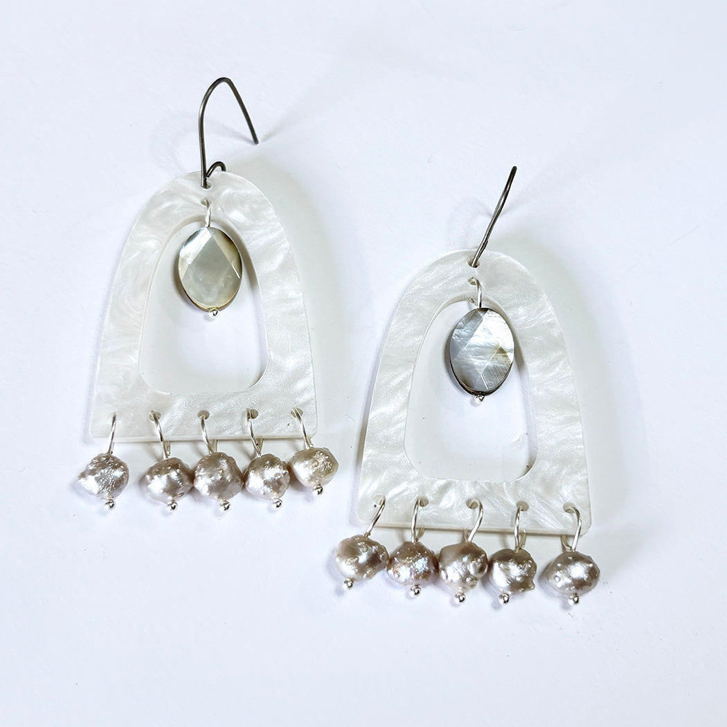 A pair of handmade dangle earrings featuring a white mother-of-pearl acrylic arch frame, a central oval faceted mother-of-pearl stone, and five small silver druzy pearl beads hanging from each earring on silver hooks.