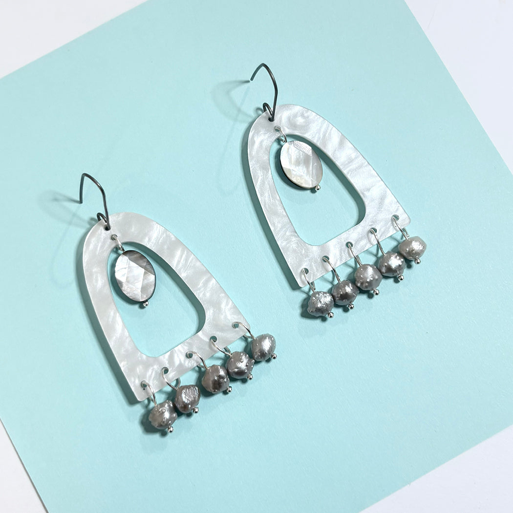 A pair of white pearl acrlic dangle earrings with open arch-shaped metal frames, oval faceted beads, and multiple small baroque pearl beads at the bottom, displayed on a light aqua background.