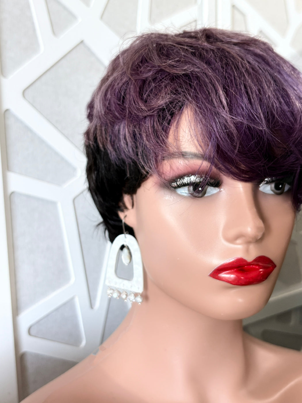 A short, textured wig with dark roots and a purple ombre top, styled on a mannequin head with dramatic makeup and white statement earrings.