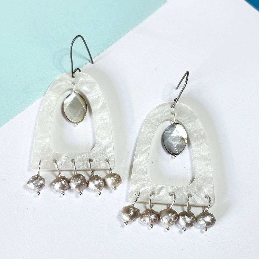 A pair of white mother or pearl acrylc dangle earrings with textured white arch-shaped pendants, oval mother-of-pearl center stones, and multiple dangling druzy pearl bead drops, displayed on a white and light blue background.