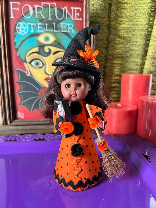 Halloween-themed doll in an orange and black outfit with a broom, framed 'Fortune Teller' sign in the background.