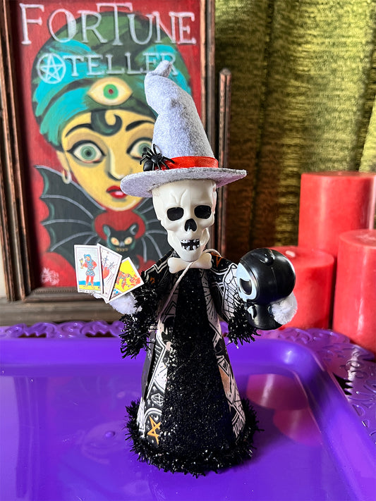 Decorative skeleton witch figure with a hat and tarot cards on a purple surface, with a colorful fortune teller poster in the background.