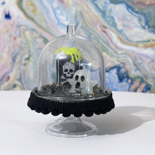 Decorative terrarium with a plastic dome featuring a tombstone, ghost, and spider on a colorful abstract background.