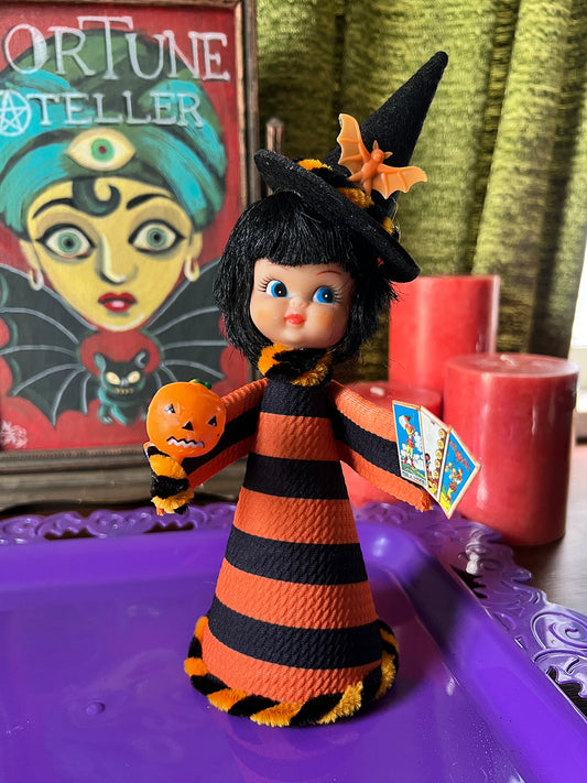 Halloween-themed figurine with striped dress and witch hat, tarot cards, and candles on a purple surface.