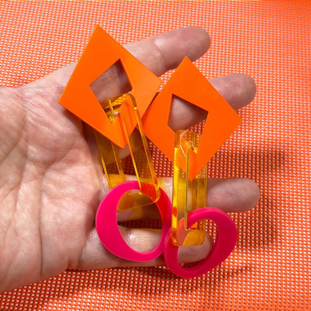 Hand holding large mismatched chunky acrylic chain earrings in orange, transparent yellow and hot pink.