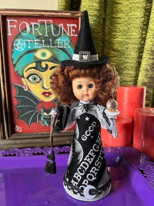 Vintage look Witch doll with a witch hat, broom and crystal ball in a black outfit in front of a 'Fortune Teller' poster.