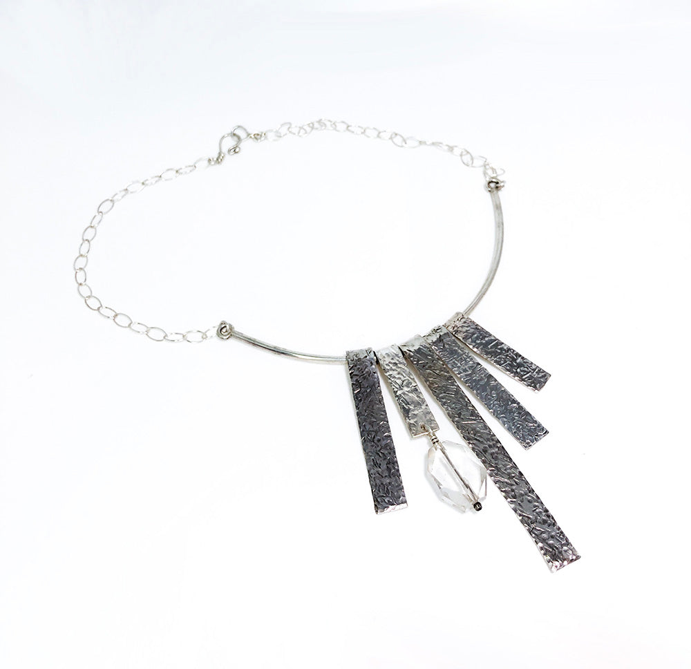Sterling silver mid-century brutalist style necklace featuring asymmetrical textured rectangular fringe bars and a faceted clear quartz crystal dropping from a sleek curved metal collar on a silver link chain. On a white background